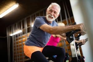 An elderly gentlemen rides a stationary bike to stay in shape. Working out can help lower Healthcare costs dramatically.