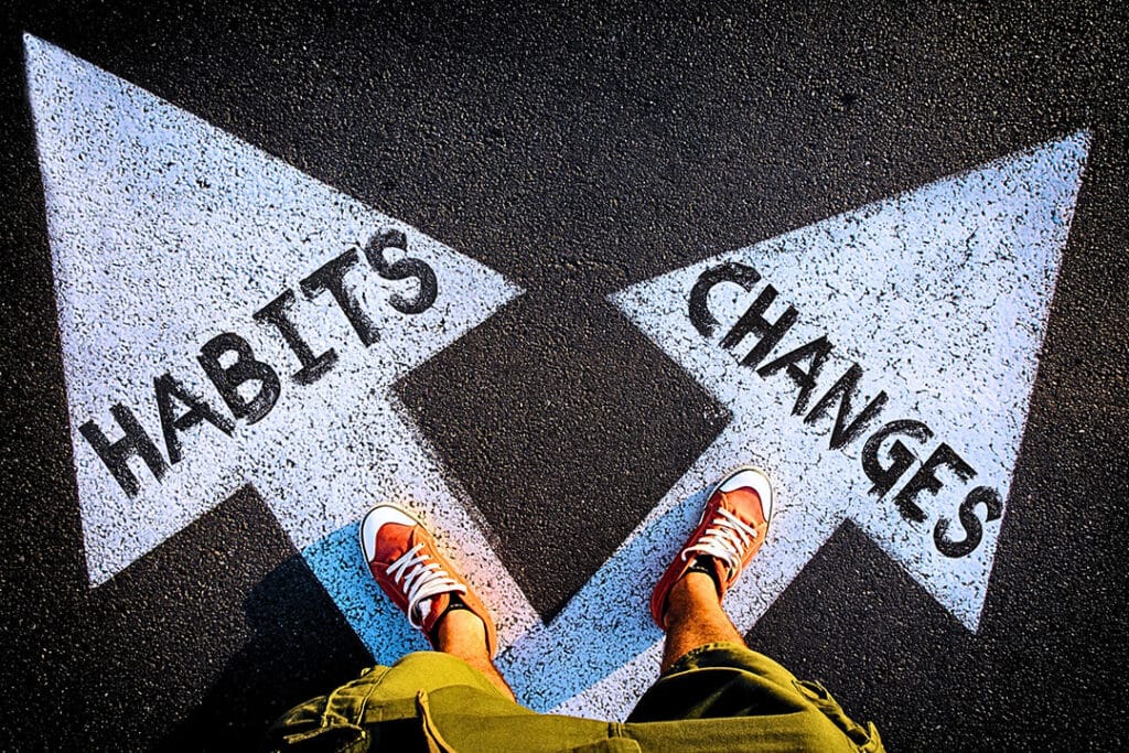 A person stands on two arrows pointing differen directions. One says "Habits" while the other says "Changes." To lower healthcare costs, many people must make change and leave old habits.