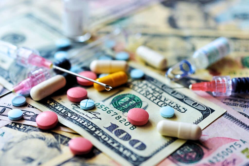 pills and insulin needles sit atop $100 bills, representing the pain of expensive prescription medications.